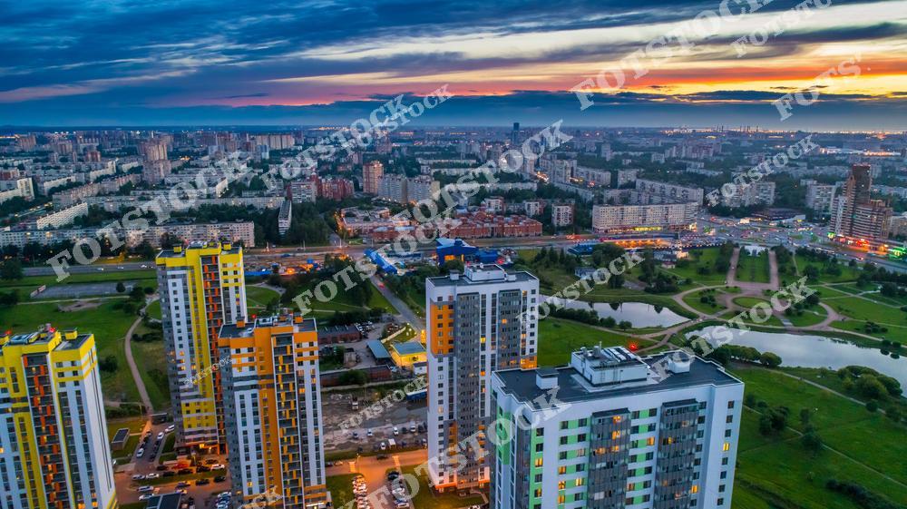 Панорамная фотография жилого квартала Купчино в Санкт-Петербурге, снятая с дрона в вечернее время. На снимке видны современные многоэтажные дома, городская инфраструктура и улицы района, освещенные мягким светом заката. Снимок подчеркивает современную архитектуру и плотную городскую застройку, а также гармоничное сочетание жилых кварталов с окружающей городской средой. Фотография отлично подходит для иллюстраций о городской жизни, недвижимости, урбанистических проектов и публикаций о современном Санкт-Петербурге. Вид с высоты раскрывает масштаб и структуру района, создавая атмосферу вечернего города и современные городские пейзажи.